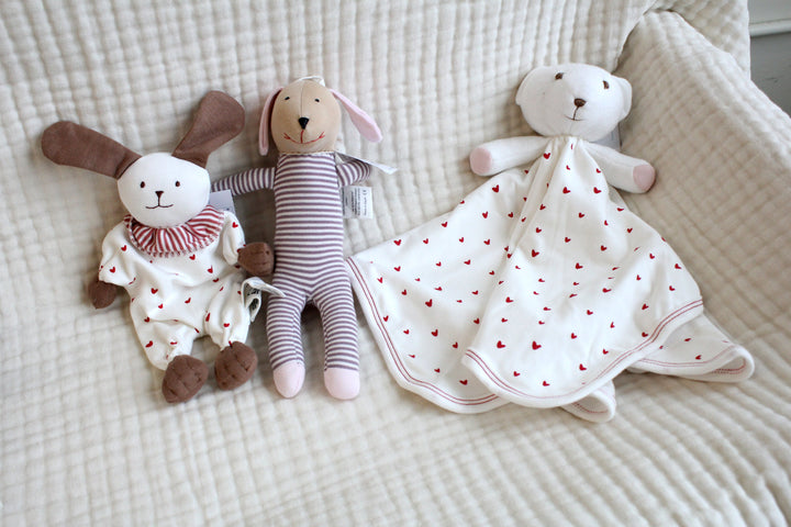 Three soft organic cotton baby toys including a puppy a bear, and a blanket with heart patterns on a textured surface.