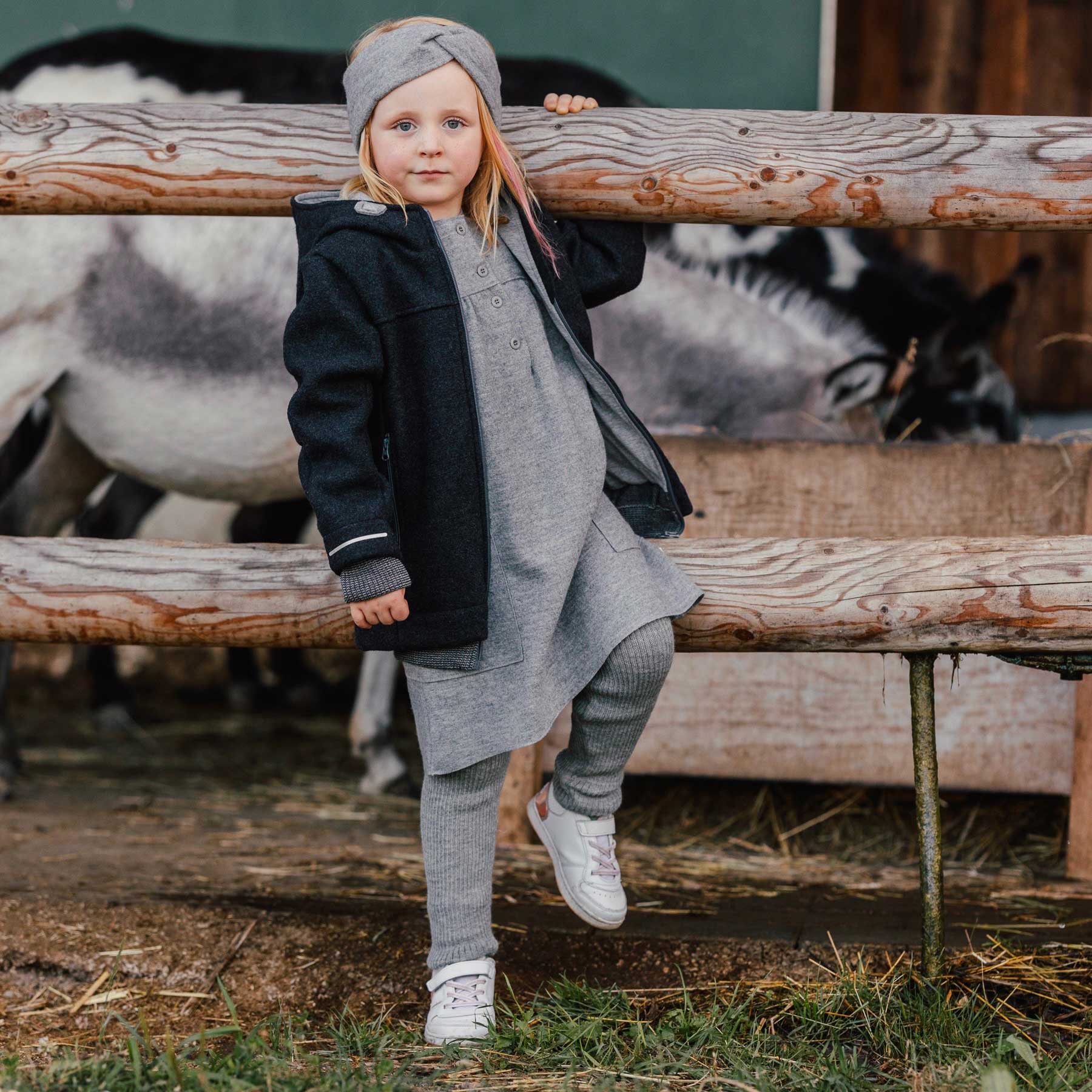 Child in gray outfit standing behind a wooden fence with animals in the background