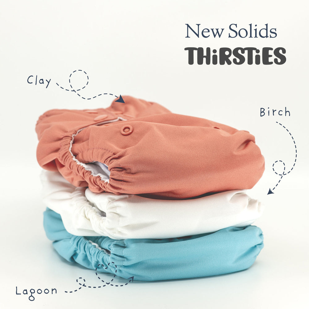 Stack of cloth diapers in clay, birch, and lagoon colors with 'New Solids Thirsties' text.