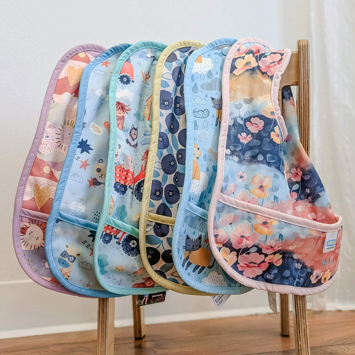 Set of colorful Thirsties baby bibs on a wooden stand against a white wall.