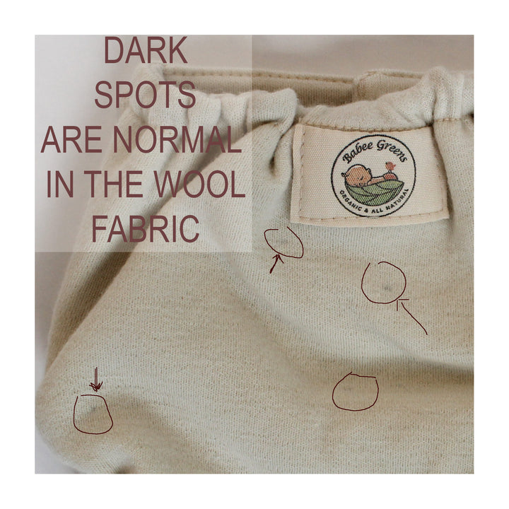 Close-up of a beige wool fabric garment with a sewn-on label reading 'Baboo Greens Organic & All Natural.' Text overlay says 'DARK SPOTS ARE NORMAL IN THE WOOL FABRIC,' with a few dark marks and hand-drawn circle arrows pointing to visible stains on the fabric.