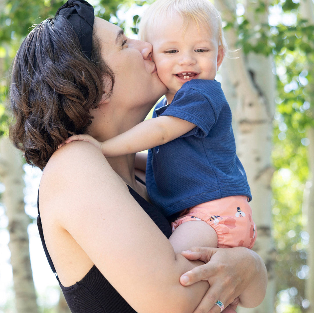 A woman outdoors holds and kisses a smiling toddler wearing a navy blue short-sleeve shirt and Thirsties Let it Be all in one cloth diaper