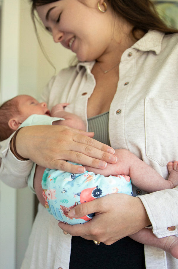 A woman holding a baby wearing a light blue and pink woodland print cloth diaper, with the woman wearing a cream-colored button-up shirt.