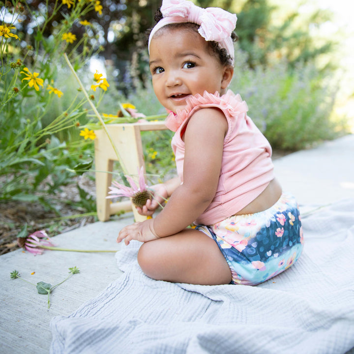 Child in a pink top and floral Thirsties Poppy print diaper sitting outdoors with flowers and greenery.