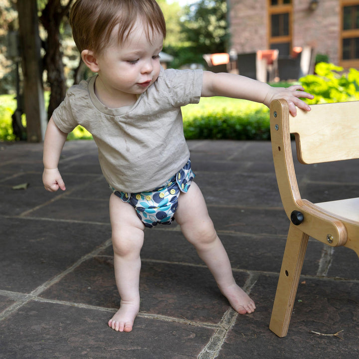 Child in a Thirsties all in one reusable  diaper  in Blueberry print standing on a stone patio with a wooden chair nearby.