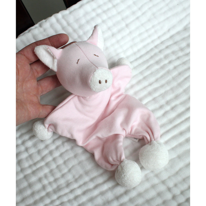 An organic cotton pink plush pig baby lovey comforter with white pom-pom feet and ears, held by a hand on a white textured blanket.