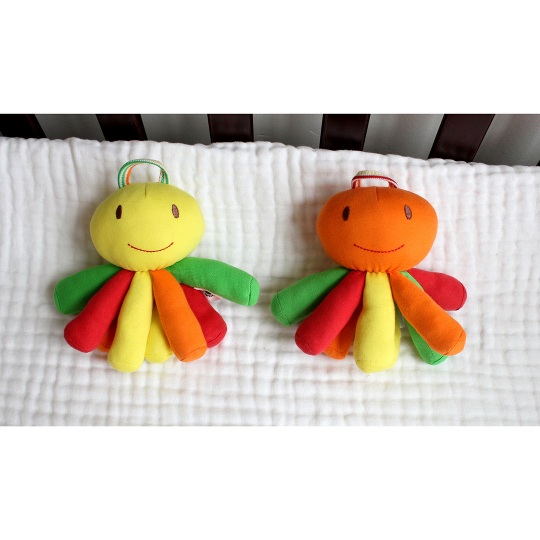 Under the Nile organic cotton baby toys Two colorful plush octopus baby toys side by side on a white textured blanket, one with a yellow head and green/red/orange tentacles, and the other with an orange head and green/red/yellow tentacles.